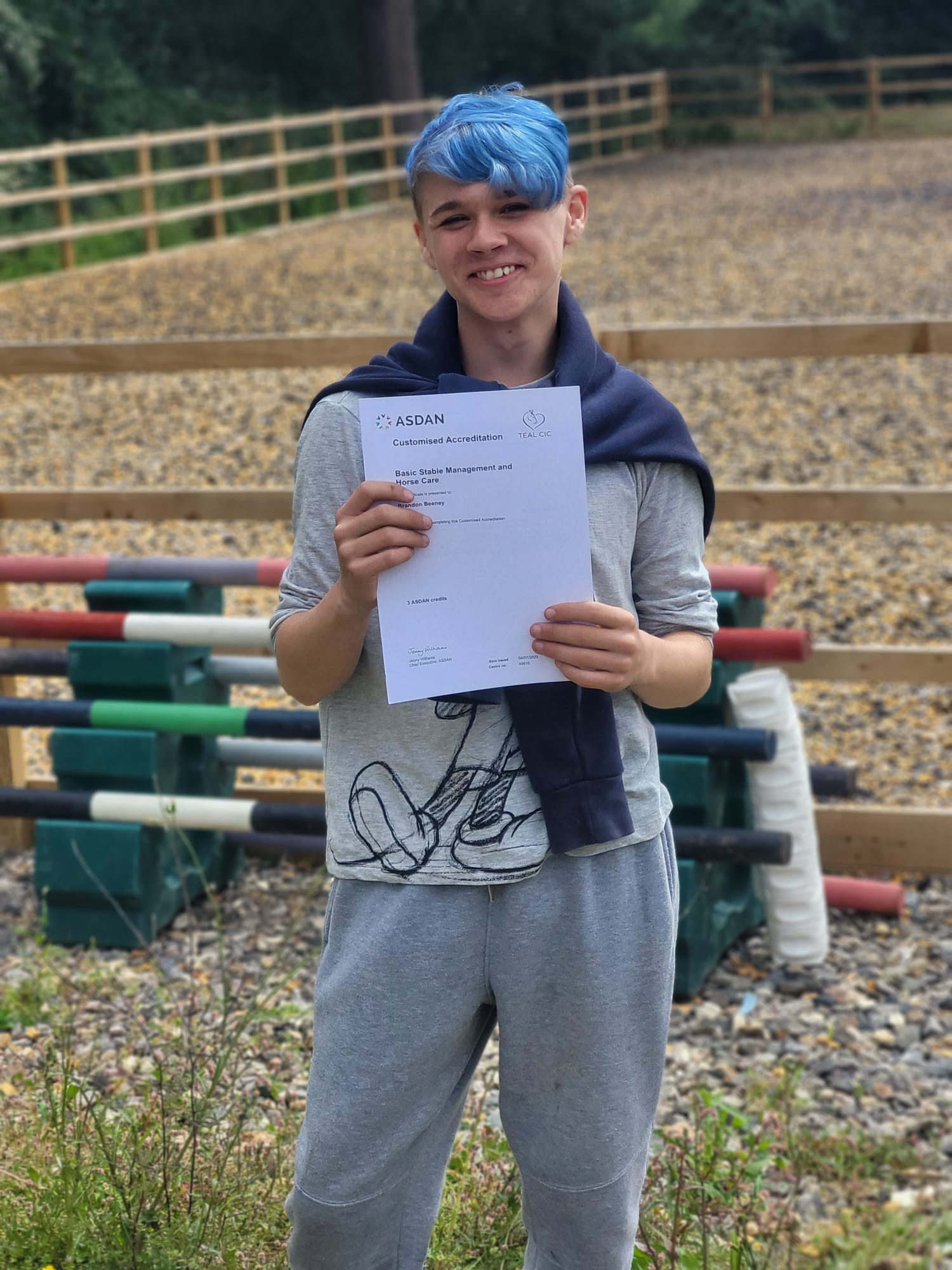 Young person with blue hair outside a stable holding up their ASDAN certificate and smiling